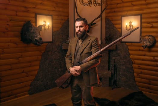 Hunter gentleman in traditional hunting clothing with old gun against fireplace. Stuffed wild animals, bear skin and other trophies on background. Old-fashioned style