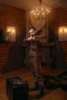 Senior hunter in retro traditional hunting clothing aims of the antique shotgun against old chest. Fireplace, stuffed wild animals, bear skin and other trophies on background