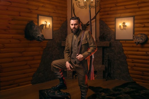 Respectable hunter man with old gun in retrro style traditional hunting clothing standing against antique chest. Retro rifle, vintage clothes. Room with fireplace, stuffed wild animals, bear skin and other trophies on background