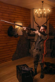 Hunter man aims of the antique hunting rifle. Fireplace, stuffed wild animals, bear skin and other trophies on background