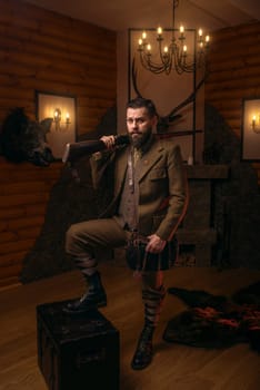 Respectable hunter man with old gun in retrro style traditional hunting clothing standing against antique chest. Retro rifle, vintage clothes. Room with fireplace, stuffed wild animals, bear skin and other trophies on background