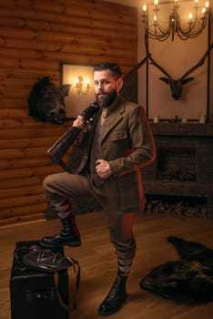 Respectable hunter man with old gun in retrro style traditional hunting clothing standing against antique chest. Retro rifle, vintage clothes. Room with fireplace, stuffed wild animals, bear skin and other trophies on background