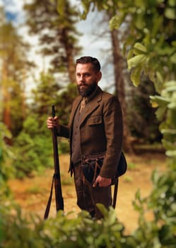 Portrait of breaded hunter man in traditional hunting clothing with old rifle, green forest on background. Hunt lifestyle