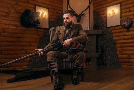 Hunter man in vintage hunting clothing sitting in a chair with antique rifle. Fireplace, stuffed wild animals, bear skin and other trophies on background. Hunt lifestyle