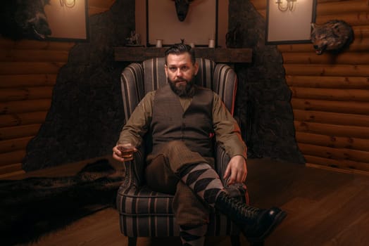 Hunter man in traditional british clothing sitting in a chair after hunting and drink whiskey. Fireplace, stuffed wild animals, bear skin and other trophies on background. Hunt lifestyle