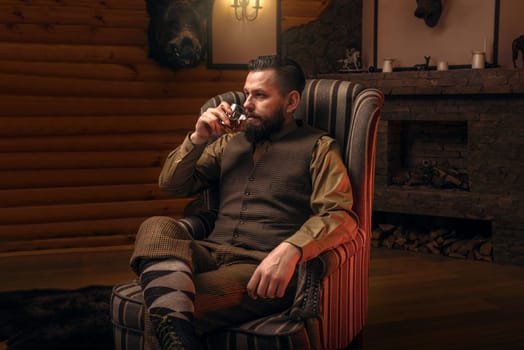 Hunter man in traditional vintage hunting clothing drink luxury alcohol after successful hunt. Fireplace, stuffed wild animals, bear skin and other trophies on background