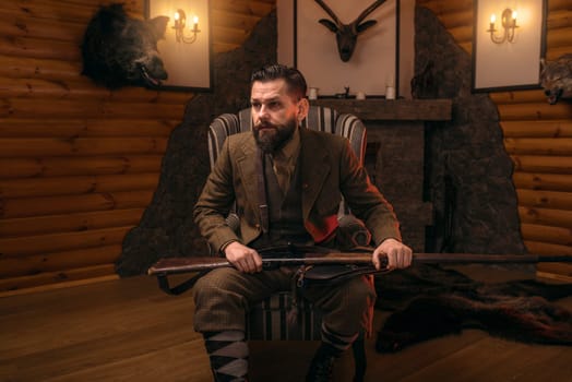 Hunter man with old gun in vintage traditional hunting clothing standing against antique chest. Fireplace, stuffed wild animals, bear skin and other trophies on background