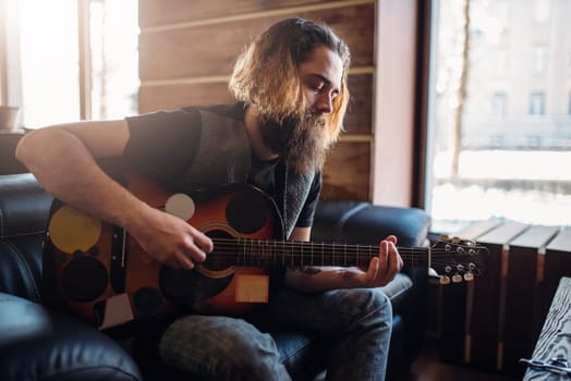 Bearded brutal man with guitar sitting on black couch. Barber shop inerior on background