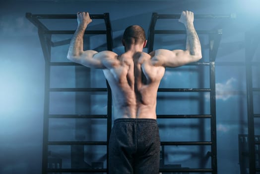 Strong sportsman training on horizontal bar in gym, back view. Muscle athlete on workout