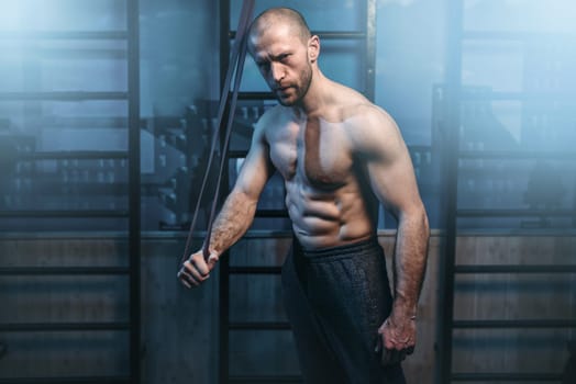 Muscular sports man with strong biceps exercises with elastic band in gym. Active fitness lifestyle. Man workout with rubber expander