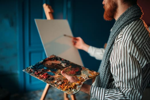 Male artist with palette and brush in hand in front of easel. Oil paint