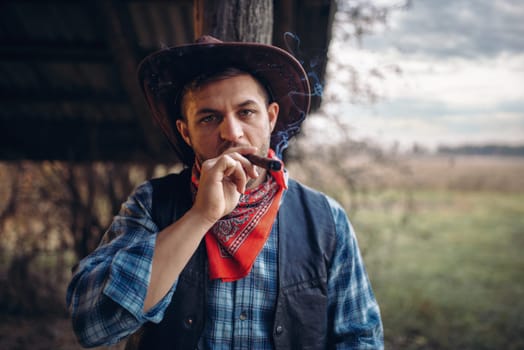 Brutal cowboy smokes a cigar, texas ranch on background, western. Vintage male person with gun on farm, wild west culture