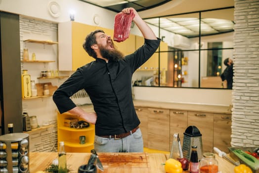 Man with opened mouth holds piece of fresh raw meat, kitchen interior on background. Chef cooking tenderloin with vegetables, spices and herbs