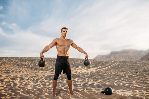 Male athlete doing exercises with two kettlebell in desert at sunny day. Strong motivation in sport, strength outdoor training
