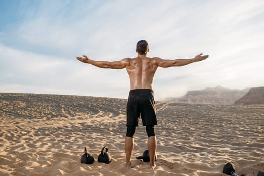 Sportsman doing exercises with weights in desert at sunny day, back view. Strong motivation in sport, strength outdoor training