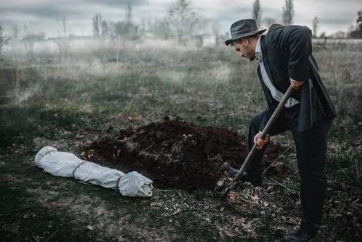 Killer is digging a grave for the victim in forest, the body wrapped in a canvas, serial maniac concept
