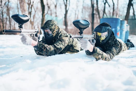 Two paintball players lies on the snow and shooting at the enemy in winter forest battle. Extreme sport game