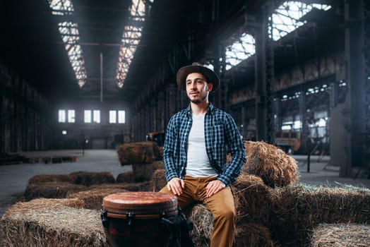 Young male drummer against african wooden drum, factory shop on background. Djembe, musical percussion instrument, bongo beats