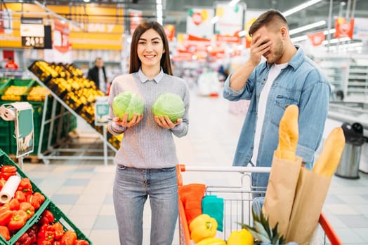 Young woman leaned the heads of cabbage to her chest, man is ashamed, funny situation in a supermarket, family shopping. Playful couple in shop, buyers in market