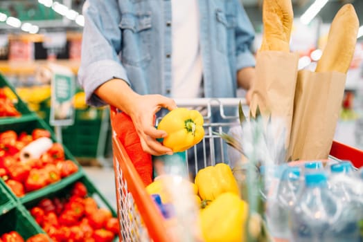 Male person puts yellow pepper in the cart with products in a supermarket, family shopping. Customer in shop, buyer in market