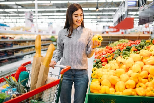Woman with cart choosing fresh sweet yellow peppers in a supermarket, family shopping. Female customer in shop, buyer in market