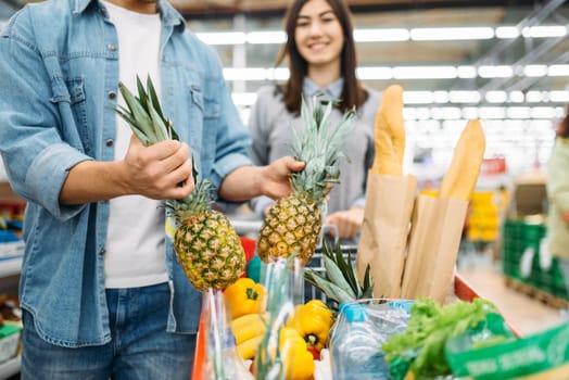 Couple puts fresh pineapples into the cart with products in a supermarket, family shopping. Customer in shop, buyer in market