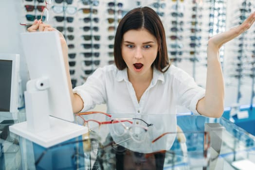 Female buyer surprised by the effect of glasses, optics store, showcase with spectacles on background. Professional eyecare in eyeglasses shop concept