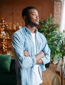 Black man with a cunning look in the living room, relaxation at home. Young african american male person standing in his house