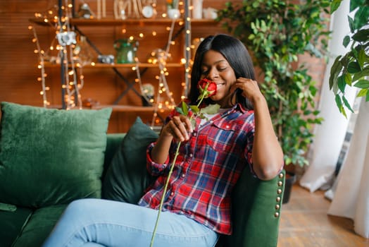 Young black woman with red rose sitting on sofa, romantic meeting at home. Happy african american female person on the couch in living room