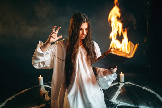 Young woman in white shirt sitting in the center of burning pentagram circle and reads spellbook, gark magic ritual