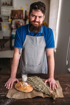 Baker against fresh baked bread bun with crispy crust, wheat bunch and glass of milk on burlap cloth. Homemade bakery concept, natural organic food.