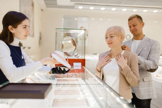 Love couple trying on gold chain. Male and female customers looking on jewels in jewelry store. Man and woman buying golden decoration. Future bride and groom in jewellery shop