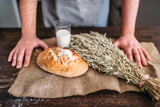 Homemade bakery concept, natural organic food. Baker hands against fresh baked bread bun with crispy crust, wheat bunch and glass of milk on burlap cloth.