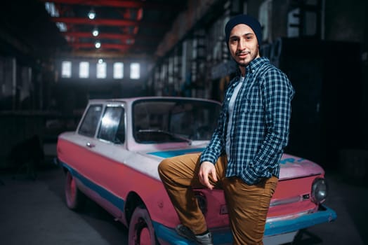 Stalker, alone traveler pose against USSR retro car, old factory interior on background. Danger zone, mysterious adventure in deserted place