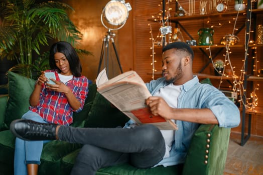 Black man reading newspaper, his woman using mobile phone on couch, relaxation at home. Happy african love couple leisures in their house