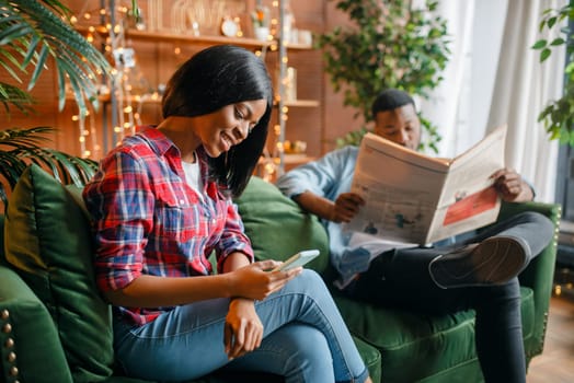 Black man reading newspaper, his woman using mobile phone on couch, relaxation at home. Happy african love couple leisures in their house
