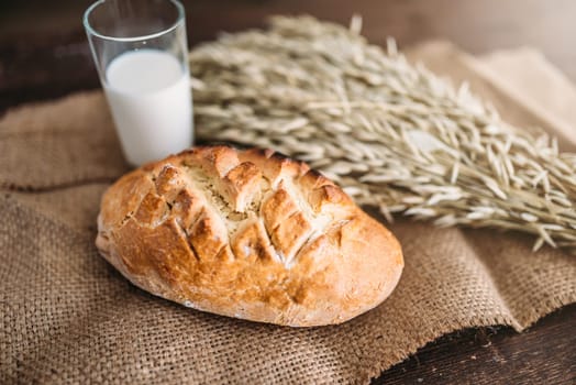Fresh bread bun with crispy crust, wheat bunch and glass of milk on burlap background, top view