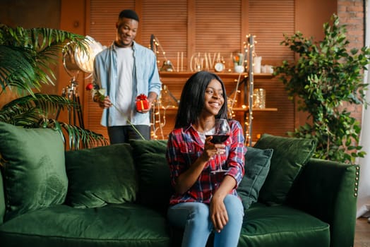 Black man with red rose and gift behind his woman with glass of wine on sofa, romantic meeting at home. Happy african love couple leisures in their house