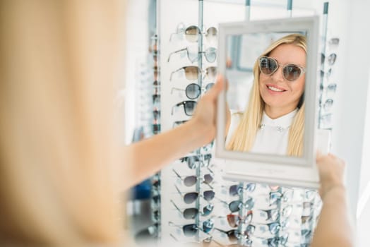 Female customer shooses sunglasses in optics store. Selection of eyes protection in glasses shop, optometry concept