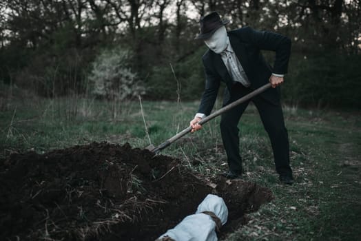 Bloody murderer is digging a grave for the victim in forest, the body wrapped in a canvas, serial maniac concept