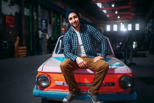 Stalker, male traveler pose against retro car, factory interior on background. Danger zone, mysterious adventure in deserted place