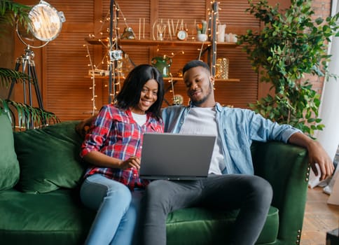 Black couple sitting on sofa and looking on laptop at home. Happy african love couple leisures in their house