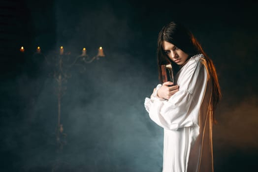 Woman in white shirt holds spellbook in hands, candles and smoke on background. Dark magic ritual, occultism and exorcism, witchcraft