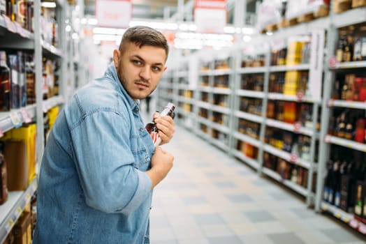 Man hides a bottle of alcohol under his shirt in supermarket. Male thief in shop, husband conducting a covert operation, shoplifling