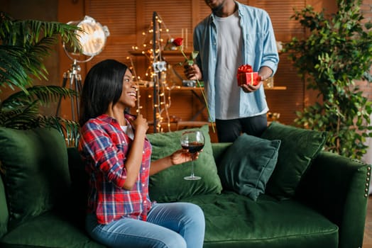 Black man with red rose and gift behind his woman with glass of wine on sofa, romantic meeting at home. Happy african love couple leisures in their house