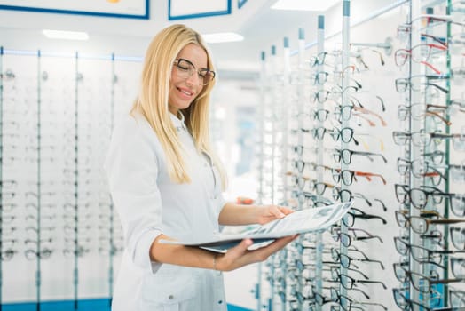Female optician with glasses catalog in hands standing against showcase with spectacles in optics store. Selection of eyeglasses with professional optometrist