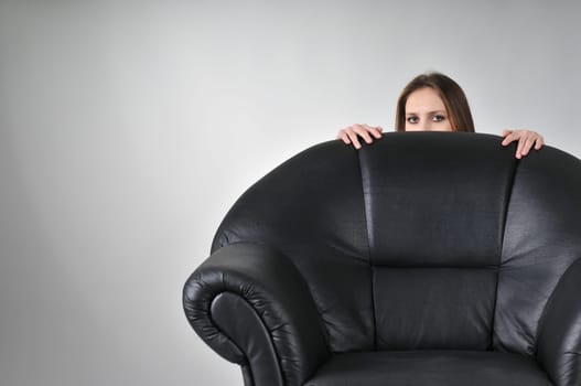 Studio portrait of young woman hidden behind big black armchair - face with hands visible