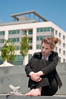 Young worried business woman siting barefoot outdoors - shoes lying next her