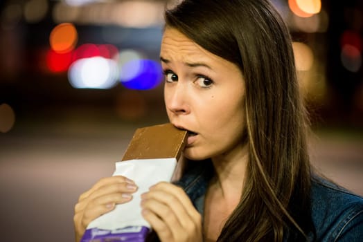 Young woman eating chocolate in street with neon lights at night