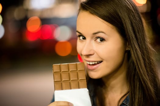 Young woman eating chocolate in street with neon lights at night